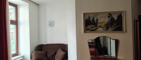 A room with a brown sofa, a wooden table, a painting of a forest, a chandelier, and a window with red curtains.