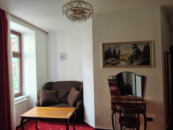 A room with a brown sofa, a wooden table, a painting of a forest, a chandelier, and a window with red curtains.