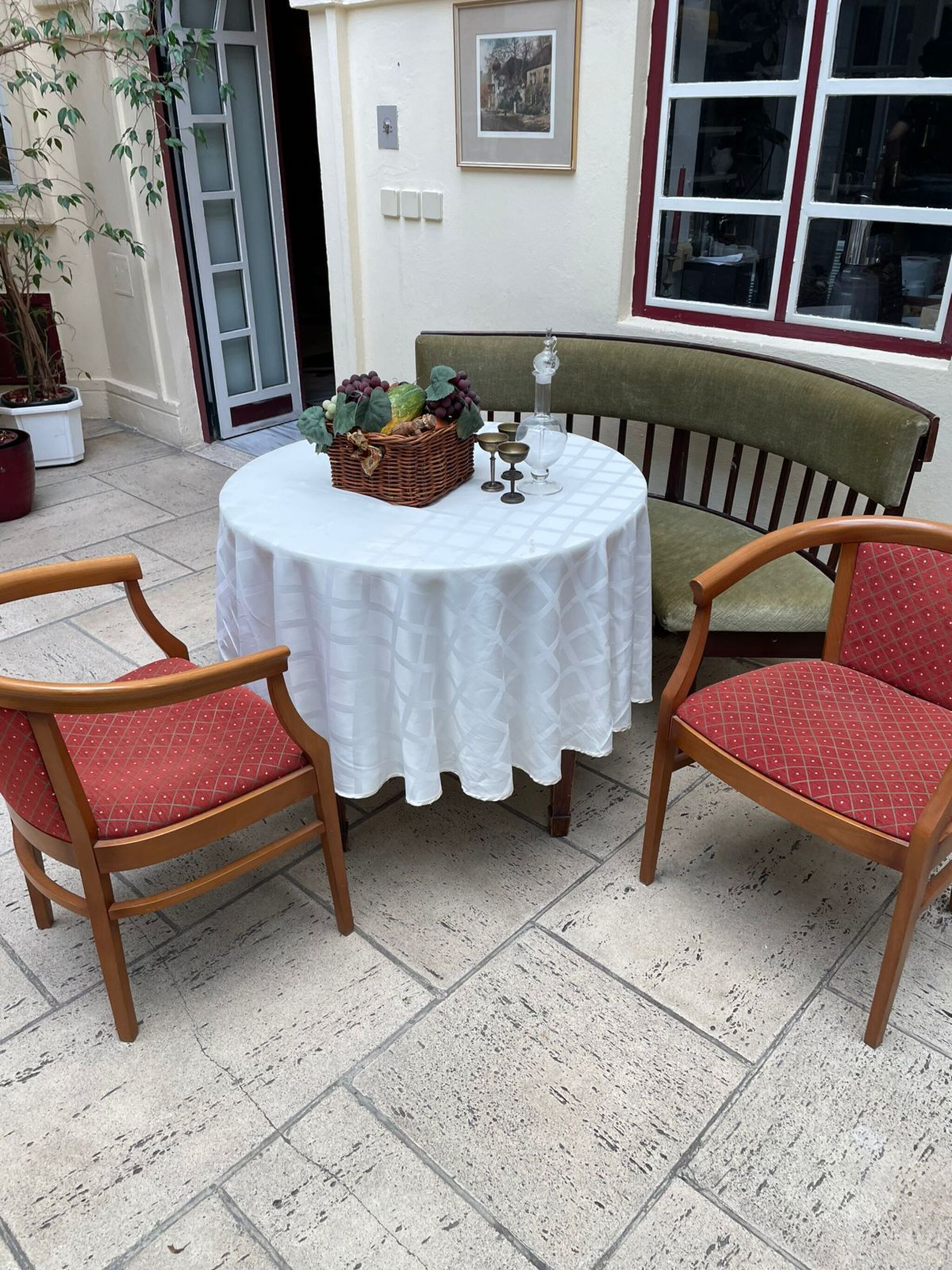 Ein runder Tisch mit weißer Tischdecke, zwei Holzstühle mit roten Kissen, ein Korbkörbchen voller Früchte und eine Glasgießkanne.