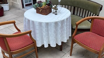 Ein runder Tisch mit weißer Tischdecke, zwei Holzstühle mit roten Kissen, ein Korbkörbchen voller Früchte und eine Glasgießkanne.