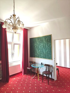 A room with a large green chalkboard, two wooden chairs, a small round table, a chandelier, and a red carpet. - Hotel Viki (Vienna)