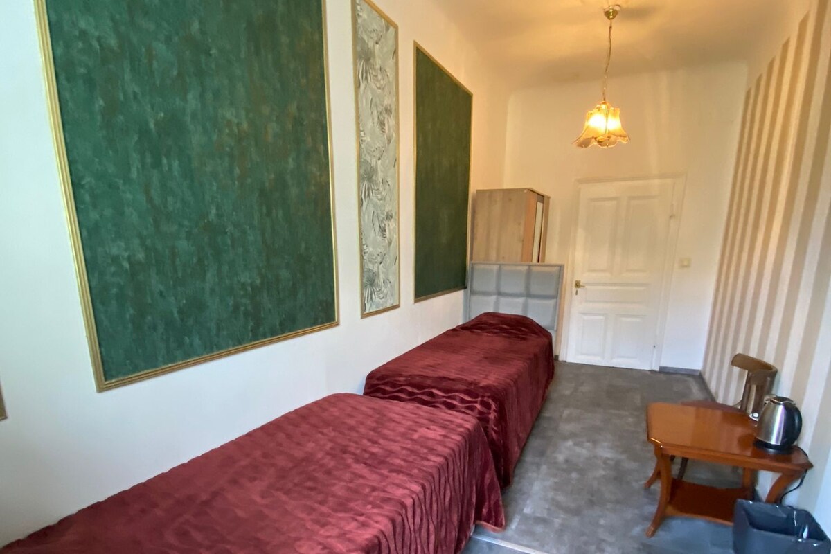 A room with two beds, a wooden table with a kettle, and a trash bin.
