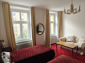 A hotel room with two beds, a sofa, a wooden table, a chandelier, and large windows with curtains. - Hotel Viki (Vienna)