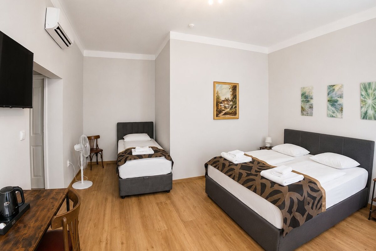 A hotel room with two beds, a desk, a chair, a fan, and artwork on the walls.