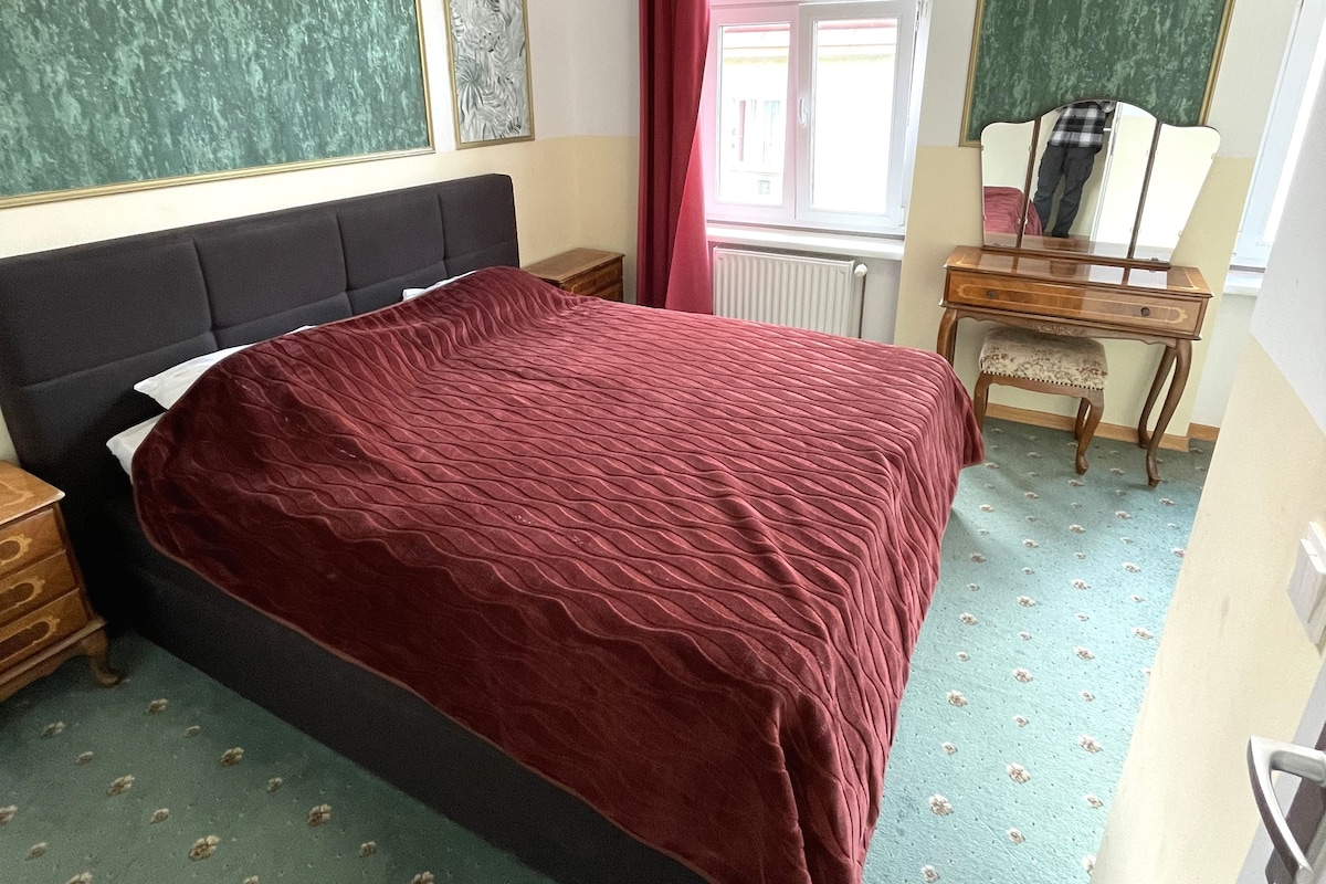A bedroom with a large bed, a wooden nightstand, a dresser, and a window with curtains.