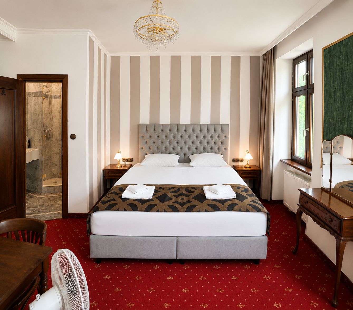 A hotel room with a large bed, two bedside tables, a mirror, a chandelier, a red carpet, and a wooden door.