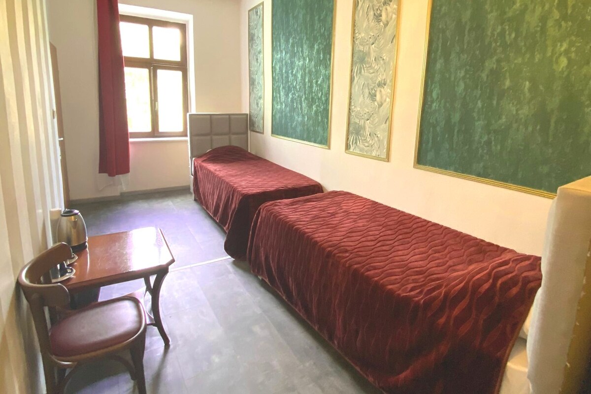 A dormitory room with two single beds, a wooden table with a chair, a radiator, and a window with a red curtain.