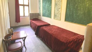 Habitación tipo dormitorio con dos camas individuales, una mesa de madera con una silla, un radiador y una ventana con cortina roja.