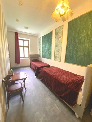 A dormitory room with two single beds, a wooden table with a chair, a radiator, and a window with a red curtain. - Hotel Viki (Vienna)