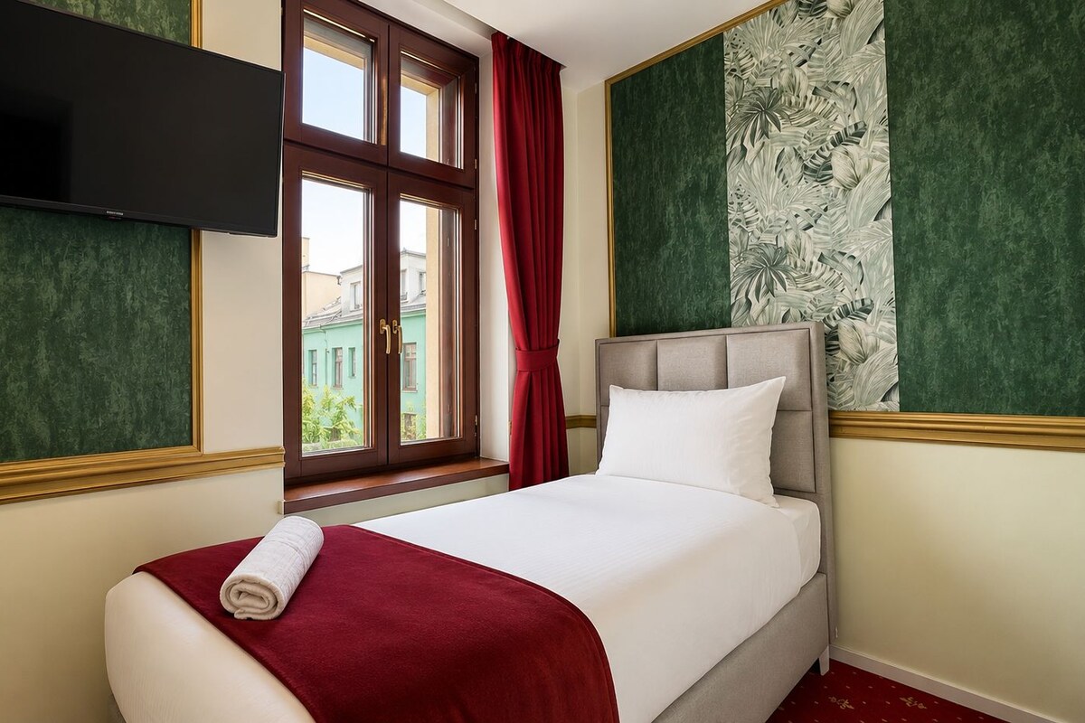 A hotel room with a bed, a flat-screen TV mounted on the wall, a window with curtains, and a green patterned wall.