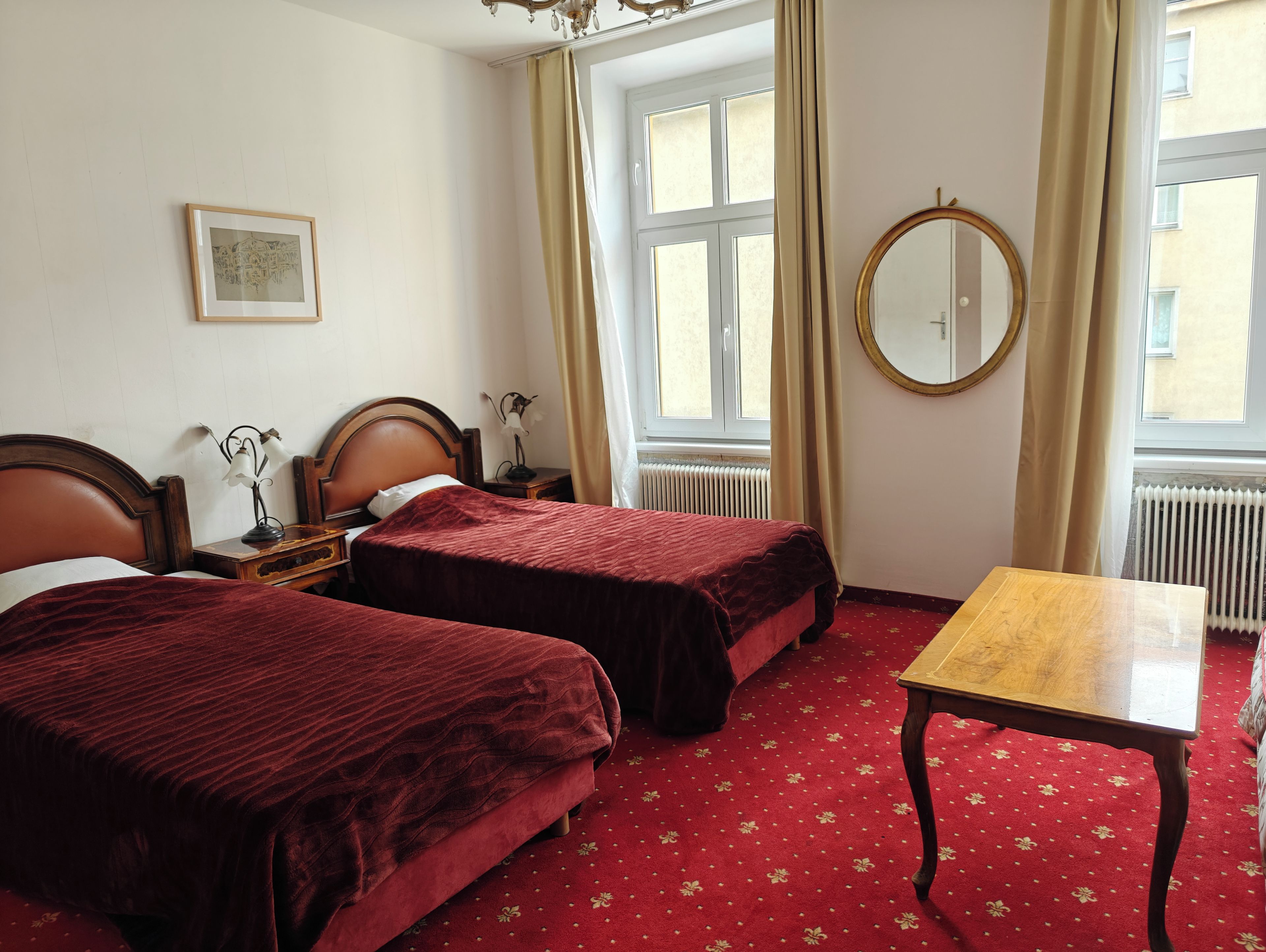 A hotel room with two beds, a wooden table, a round mirror, and a window with curtains.