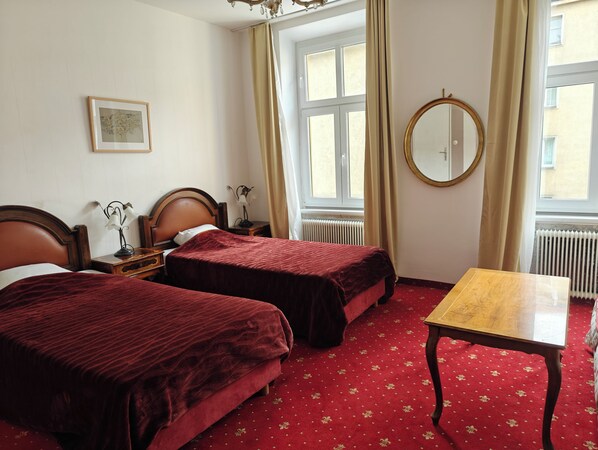 A hotel room with two beds, a wooden table, a round mirror, and a window with curtains. - Hotel Viki (Vienna)