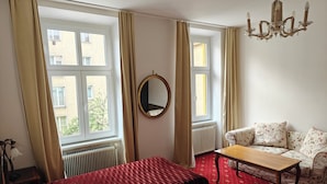 A hotel room with two beds, a sofa, a wooden table, a chandelier, and large windows with curtains. - Hotel Viki (Vienna)