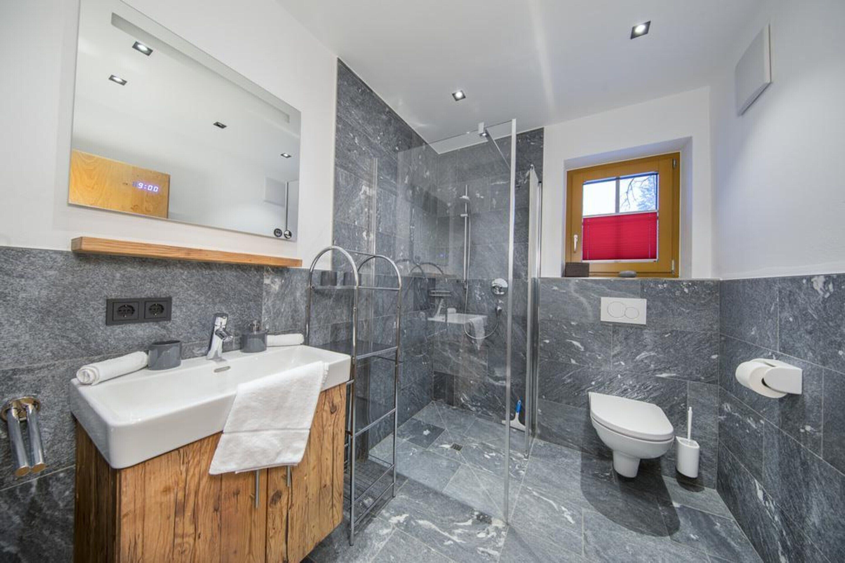 Une salle de bain moderne avec une douche en marbre, un meuble-lavabo en bois, des toilettes et une fenêtre rouge.