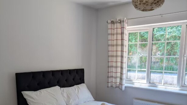 Ein ordentlich bezogenes Bett mit weißen Leinen und einem aufgerollten Handtuch, ein schwarzes, mit Stoff bezogenes Kopfteil und ein Fenster mit Karottenvorhängen.