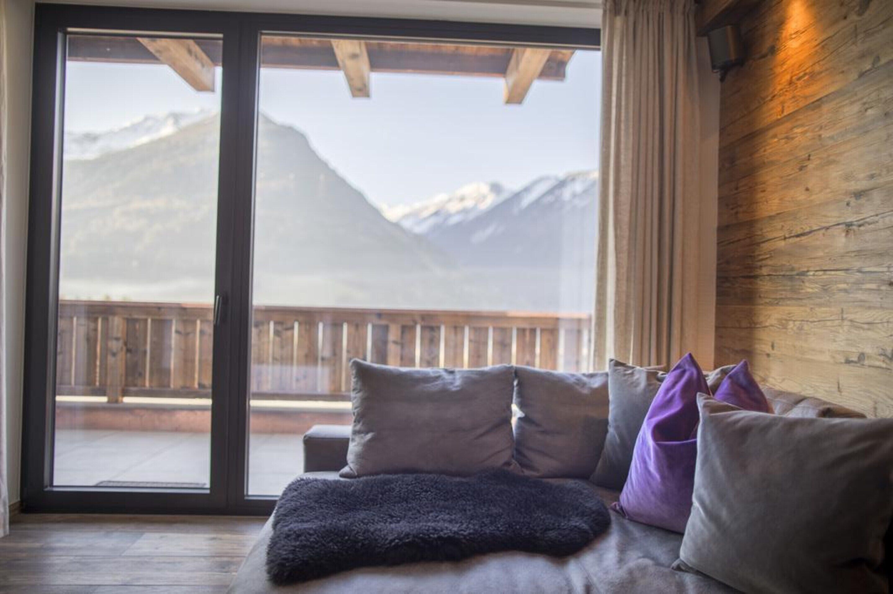 A room with a large sliding glass door leading to a balcony with a wooden railing, offering a view of snow-capped mountains.