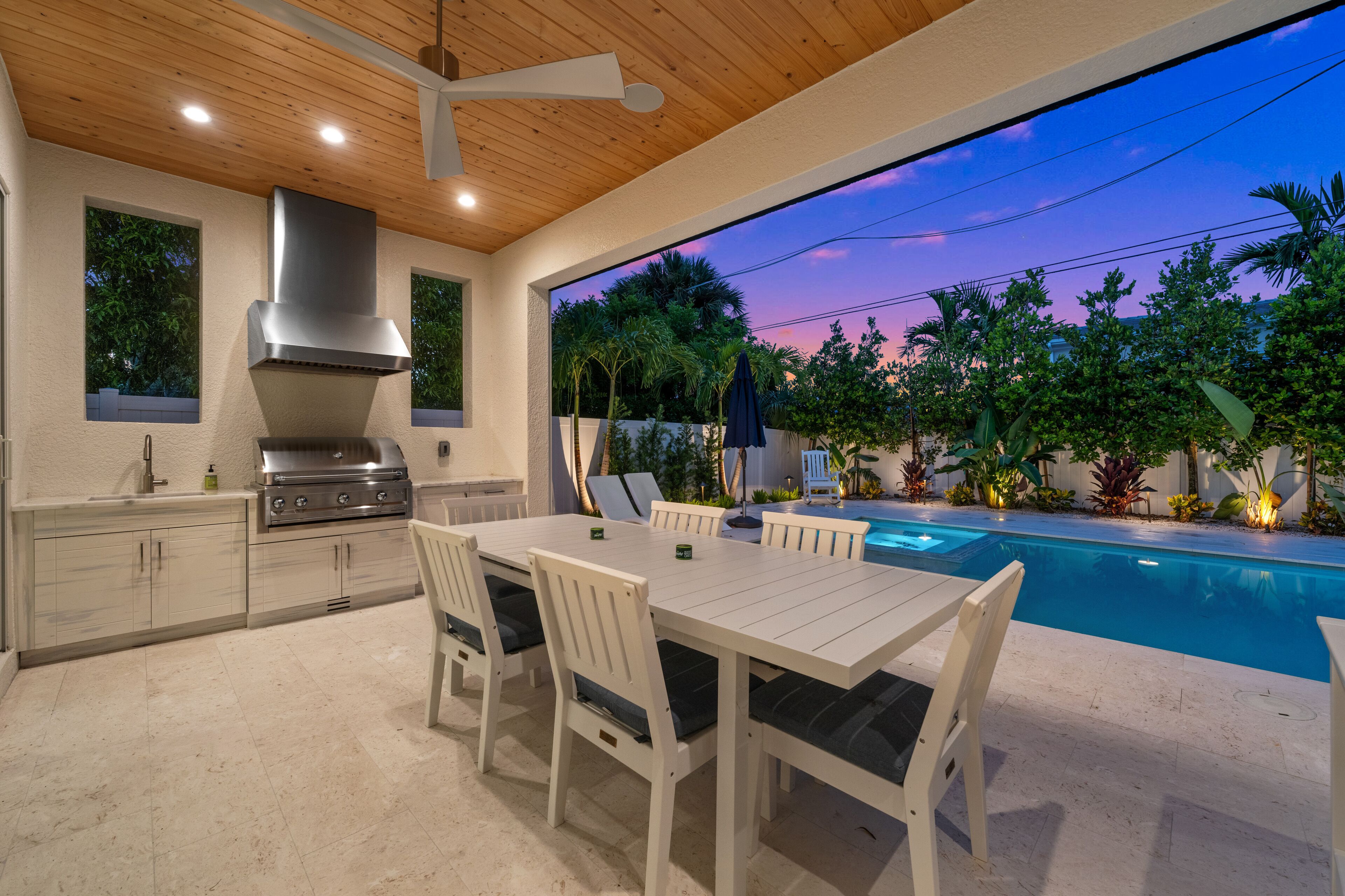 Une cuisine extérieure équipée d’un barbecue, d’une table à manger et de chaises, avec vue sur une piscine entourée d’une végétation luxuriante et sur un ciel au coucher du soleil.