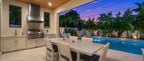 Une cuisine extérieure équipée d’un barbecue, d’une table à manger et de chaises, avec vue sur une piscine entourée d’une végétation luxuriante et sur un ciel au coucher du soleil.