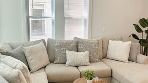 A modern living room with a sectional sofa, a wooden coffee table, and a large window with blinds. - Charming 3-bedroom House - Two Blocks from the Beach (Ventnor City)