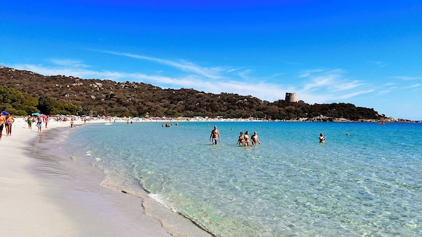 A beach with clear water, people swimming, and a castle-like structure on a hill. - Costa Rei - House 500 meters from the sea, climate, sea view terrace and garden  (Costa Rei)
