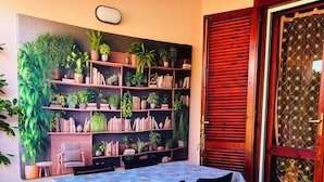 A dining area with a table set for four, surrounded by a bookshelf filled with books and plants, and a red door with a decorative glass panel. - Costa Rei - House 500 meters from the sea, climate, sea view terrace and garden  (Costa Rei)