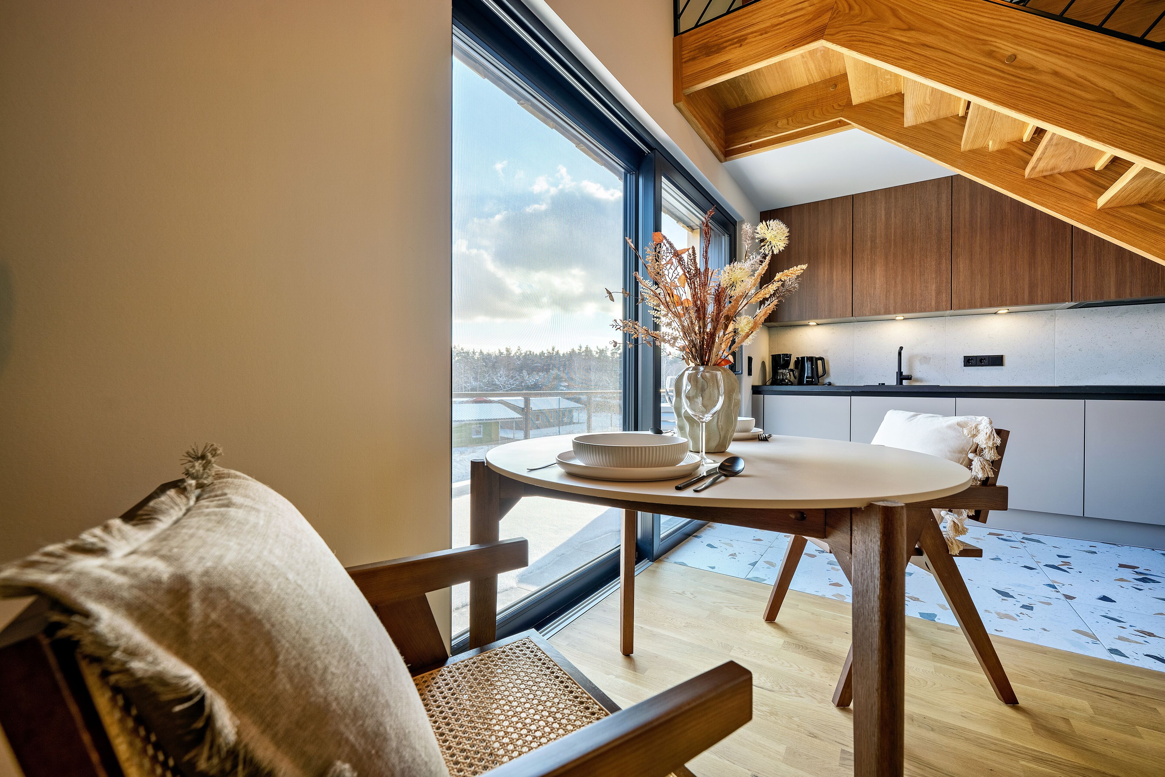 Une cuisine moderne avec des meubles en bois, un coin repas avec une table ronde et des chaises, et une grande fenêtre donnant sur l’extérieur.