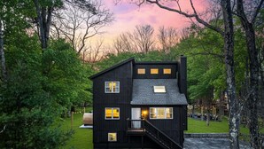 A two-story black house with a staircase leading to the entrance, surrounded by trees and a well-maintained lawn. - Escape to the Poconos | Hot Tub, Sauna, Mini-Golf (East Stroudsburg)
