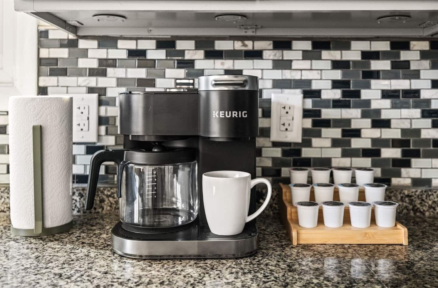 Une machine à café Keurig posée sur un comptoir en granit, avec un distributeur d’essuie-tout, une tasse blanche et un plateau rempli de gobelets à café jetables.