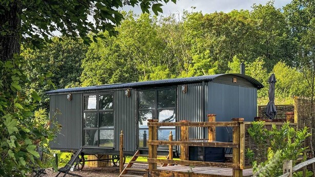 A compact, modern cabin with a metal exterior, a wooden deck, and a chimney. - The Drey, a Stunning Cumbrian Shepherd’s Hut (Carnforth)