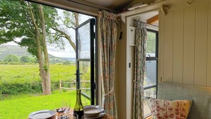 A dining table set with plates, glasses, and a bottle, with a view of a green landscape through open windows. - The Holt, a Stunning Cumbrian Shepherd’s Hut (Carnforth)