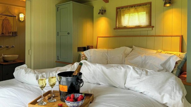 A hotel room with a bed, a bedside table with a bottle of champagne, glasses, and a tray with strawberries. - The Holt, a Stunning Cumbrian Shepherd’s Hut (Carnforth)