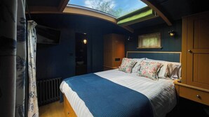 A bedroom with a skylight, a bed with floral pillows, a wooden nightstand, and a wardrobe. - The Sett, a Stunning Shepherd’s Hut & Hot Tub (Carnforth)