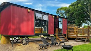 A red metal building with large windows, a deck with lounge chairs, and a grill. - The Sett, a Stunning Shepherd’s Hut & Hot Tub (Carnforth)