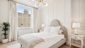 Ein Schlafzimmer mit einem großen Bett, zwei Nachttischen, einer Topfpflanze und Blick auf ein Ziegelgebäude durch das Fenster.