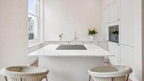 Eine moderne Küche mit weißen Schränken, einer zentralen Kochinsel, zwei Korbstühlen und Blick auf die Stadt durch ein großes Fenster.