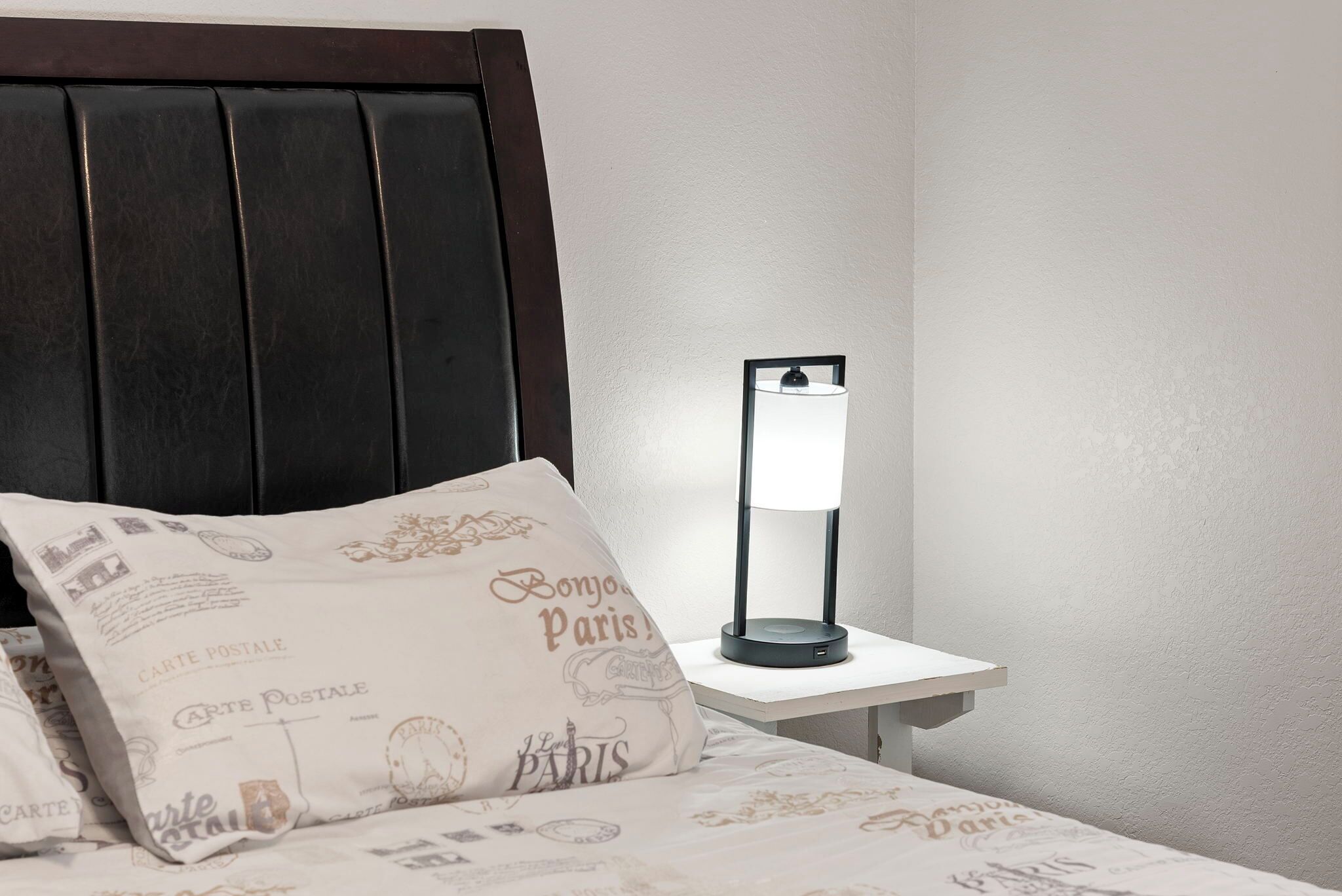A bed with a dark headboard, a white bedside table, a lamp, and a pillow with 'Carte Postale Paris' text.