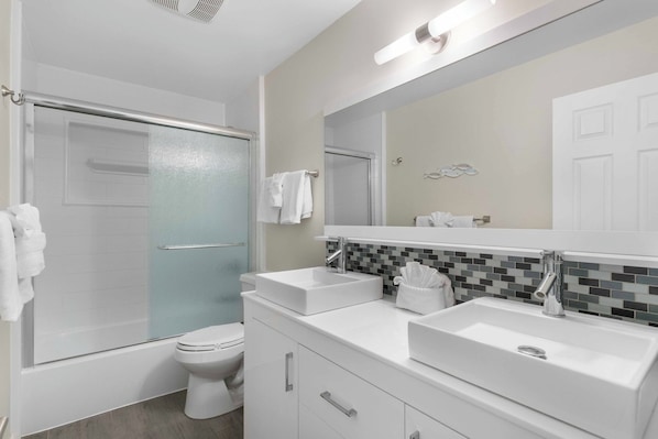Un baño moderno con ducha envuelta en vidrio, inodoro blanco y tocador con dos lavamanos y un espejo.
