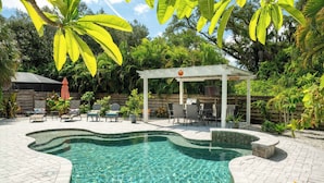 Ein Pool mit gewölbter Kante, umgeben von einer gepflasterten Terrasse mit Stühlen und einem überdachten Sitzbereich.