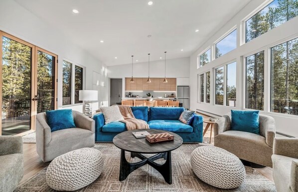 A spacious living room with a large sofa, two ottomans, and a coffee table. A kitchen is visible in the background with wooden cabinets and pendant lights.