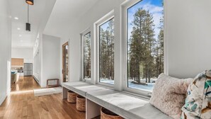 Un interior moderno con un banco junto a una ventana que da a un bosque, suelo de madera y una cocina al fondo.