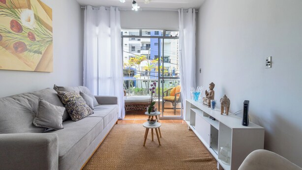 A modern living room with a grey sofa, a small white side table, and a white cabinet. A large window with white curtains provides a view of the outside. - Condomínio Praia de Taormina (Guaruja)