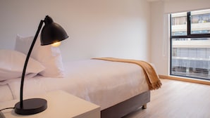 Une chambre à coucher moderne avec un lit, une lampe de chevet et une fenêtre donnant sur des immeubles.