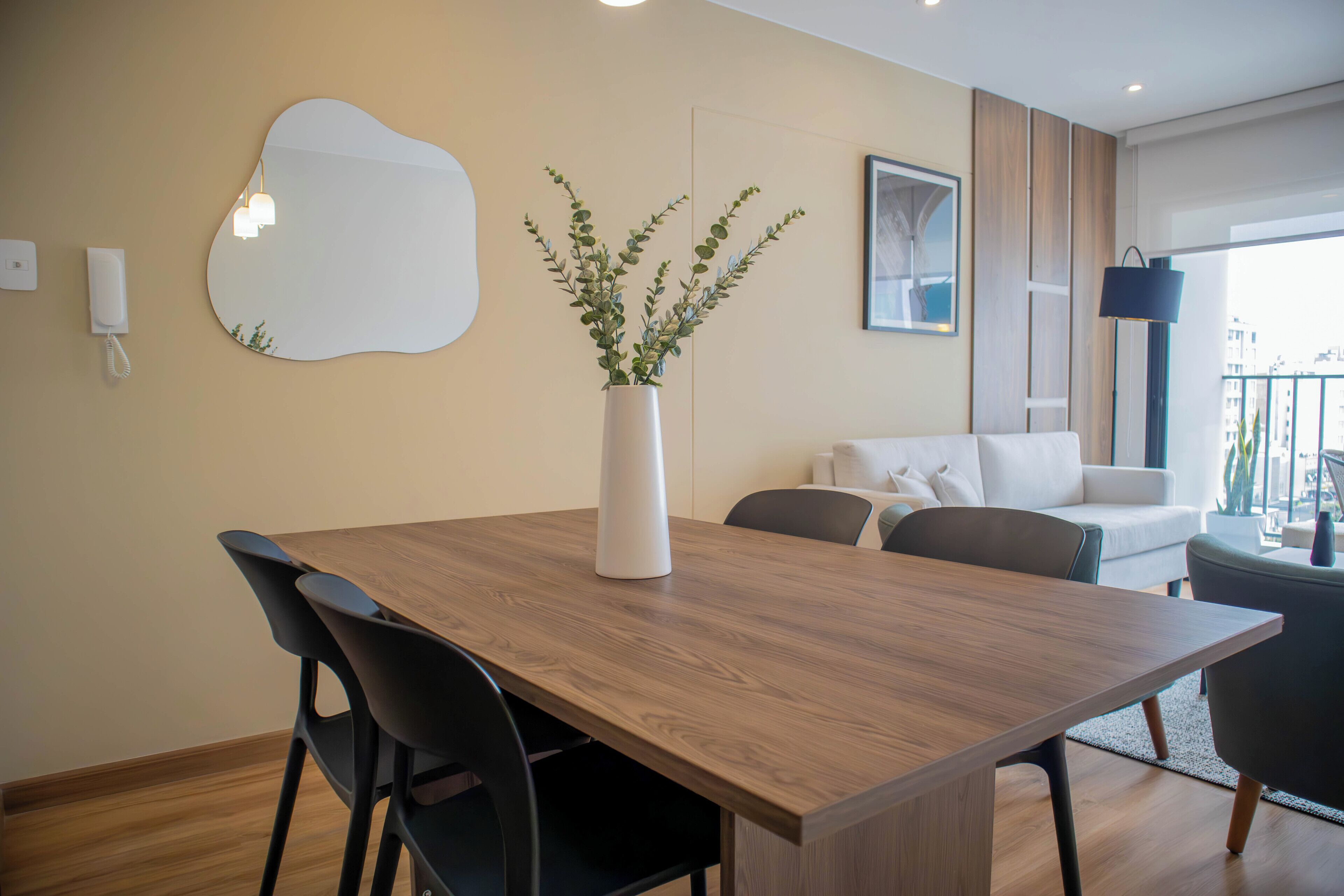 A modern living room with a wooden dining table, black chairs, a white sofa, and a large mirror on the wall.