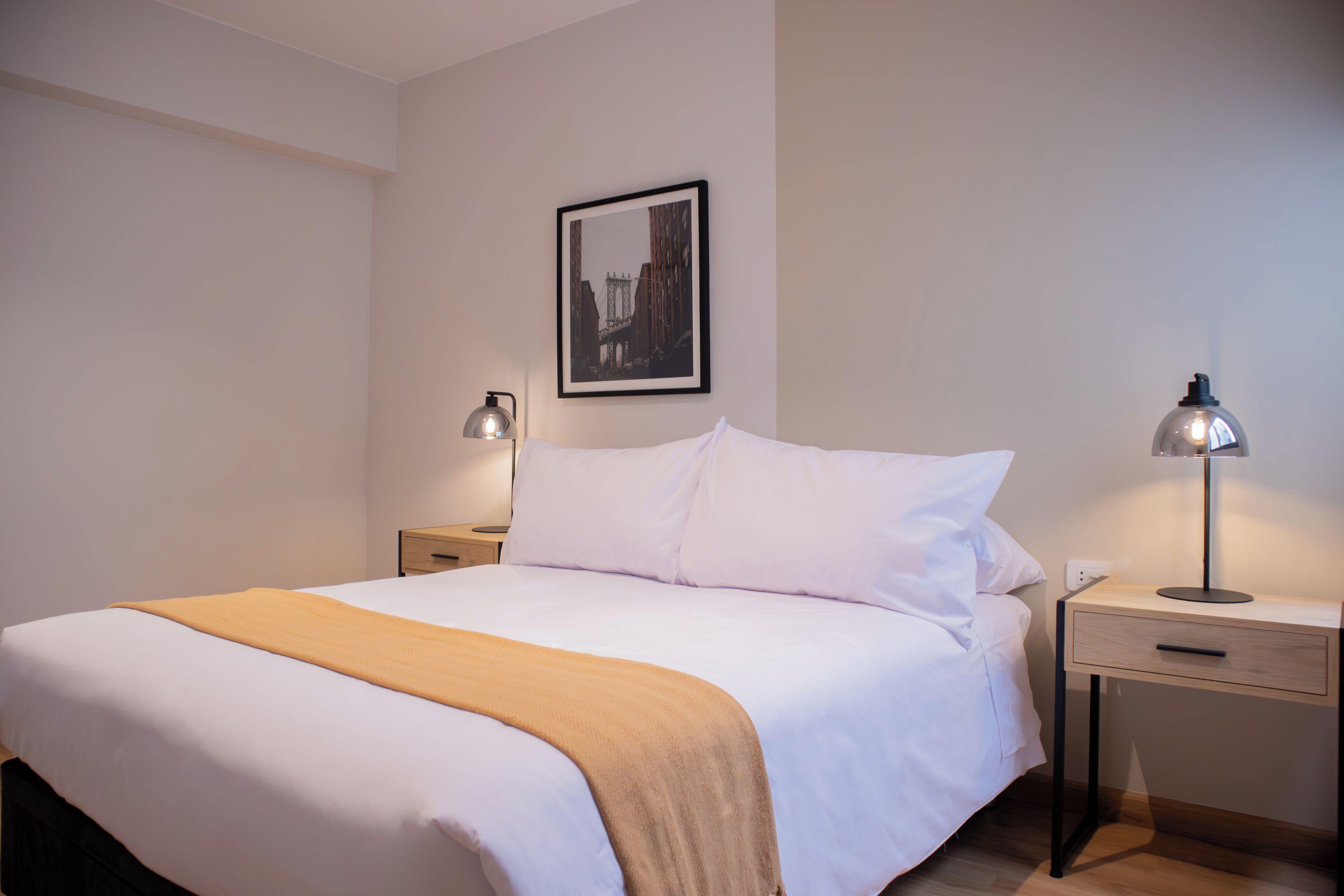 Una cama bien hecha con sábanas blancas y un edredón beige, una mesita de noche con una lámpara y un cuadro en la pared.