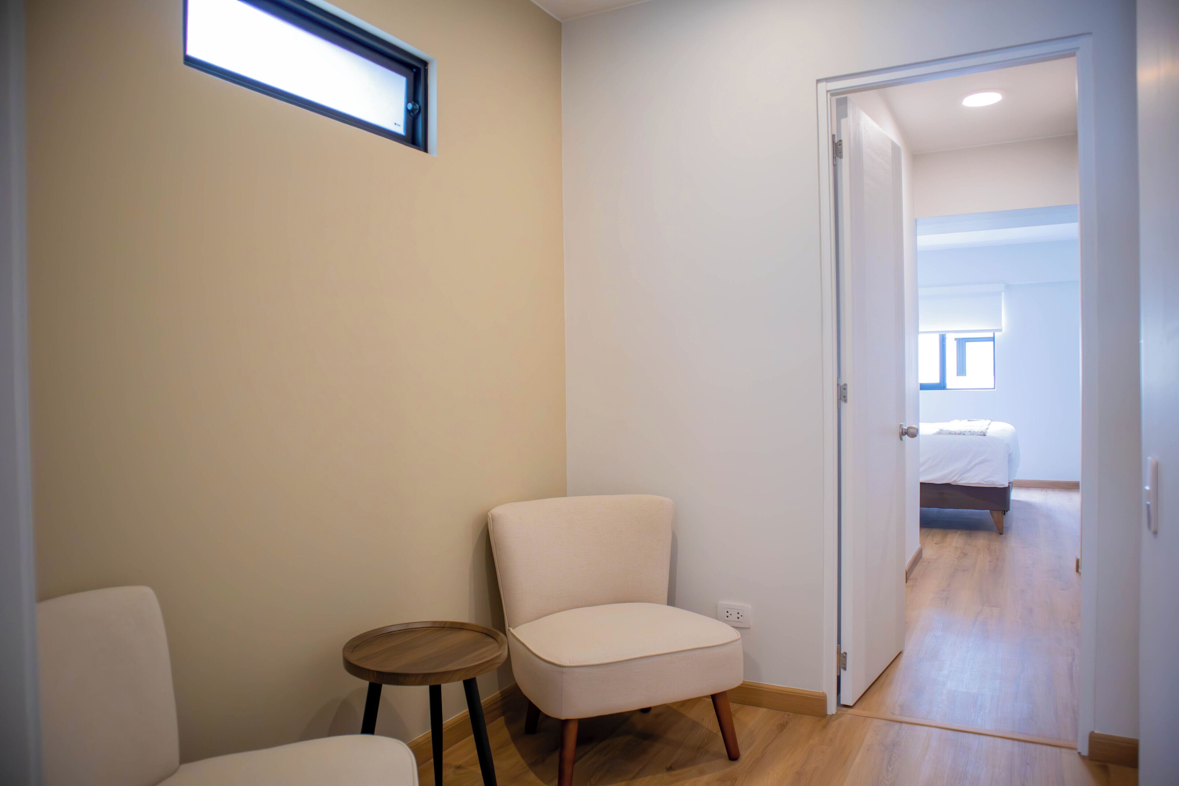 Una habitación con una silla blanca, una mesita redonda pequeña y una puerta que da a otra habitación con una cama.