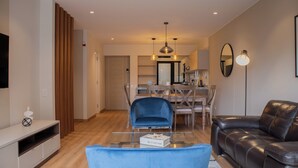 Un salon moderne avec un fauteuil bleu, un canapé en cuir noir, une table basse en verre et un coin repas comprenant des chaises en bois et un lustre.
