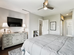A bedroom with a bed, a television, a ceiling fan, a dresser, and a door.