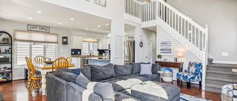 A spacious living area with a sectional sofa, a dining table with chairs, a staircase, and a kitchen in the background.