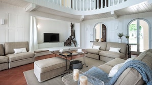 A spacious living room with a high ceiling, white walls, and a balcony. There is a large flat-screen TV, a coffee table, and multiple seating areas. - Marina dei Greci (Giardini Naxos)