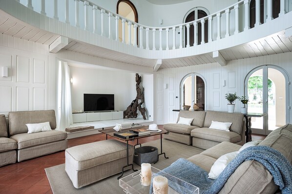 A spacious living room with a high ceiling, white walls, and a balcony. There is a large flat-screen TV, a coffee table, and multiple seating areas. - Marina dei Greci (Giardini Naxos)